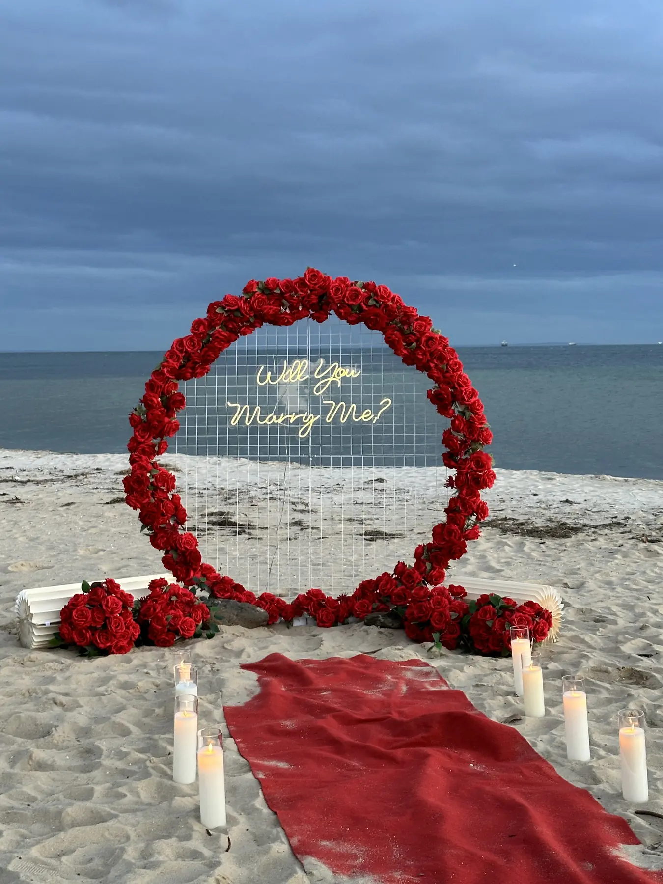 Red rose ring arch with a glowing Will You Marry Me neon sign on the sand at dusk, a red carpet aisle lined with pillar candles and the bay behind